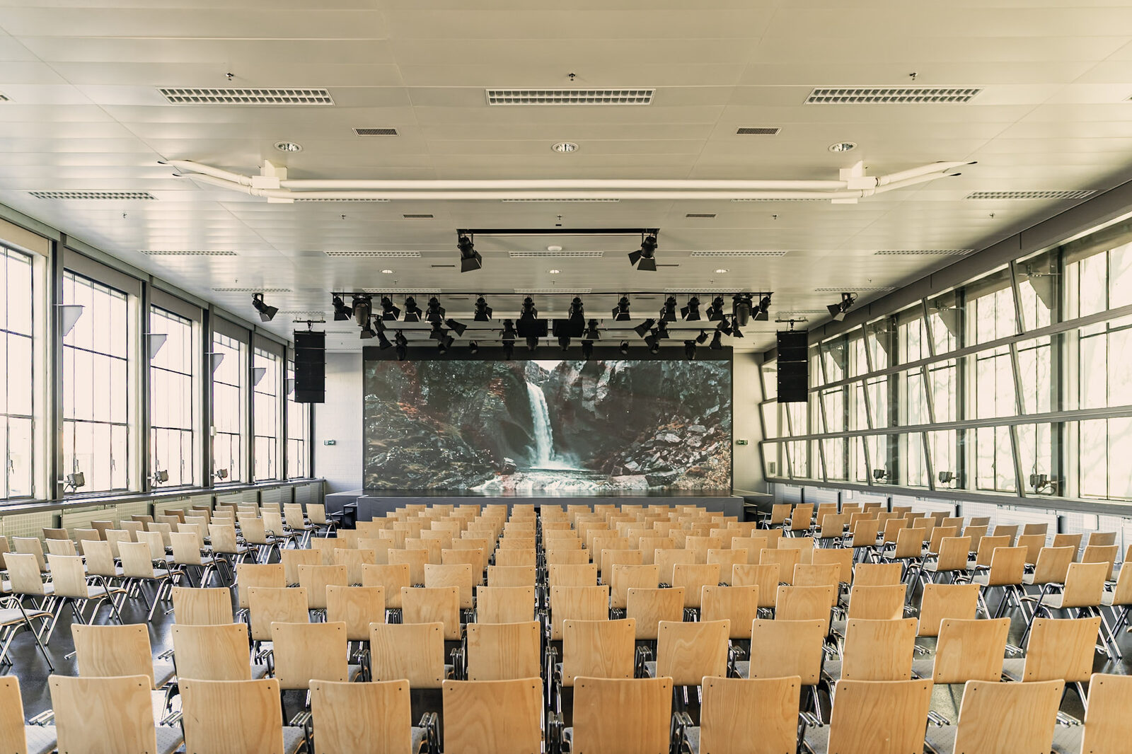 Plenarsaal der Eventfabrik Bern mit laufender LED-Wand: ein Wasserfall-Motiv auf der Bühnenwand, davor rund 300 Stühle, seitlich raumhohe Fensterfronten