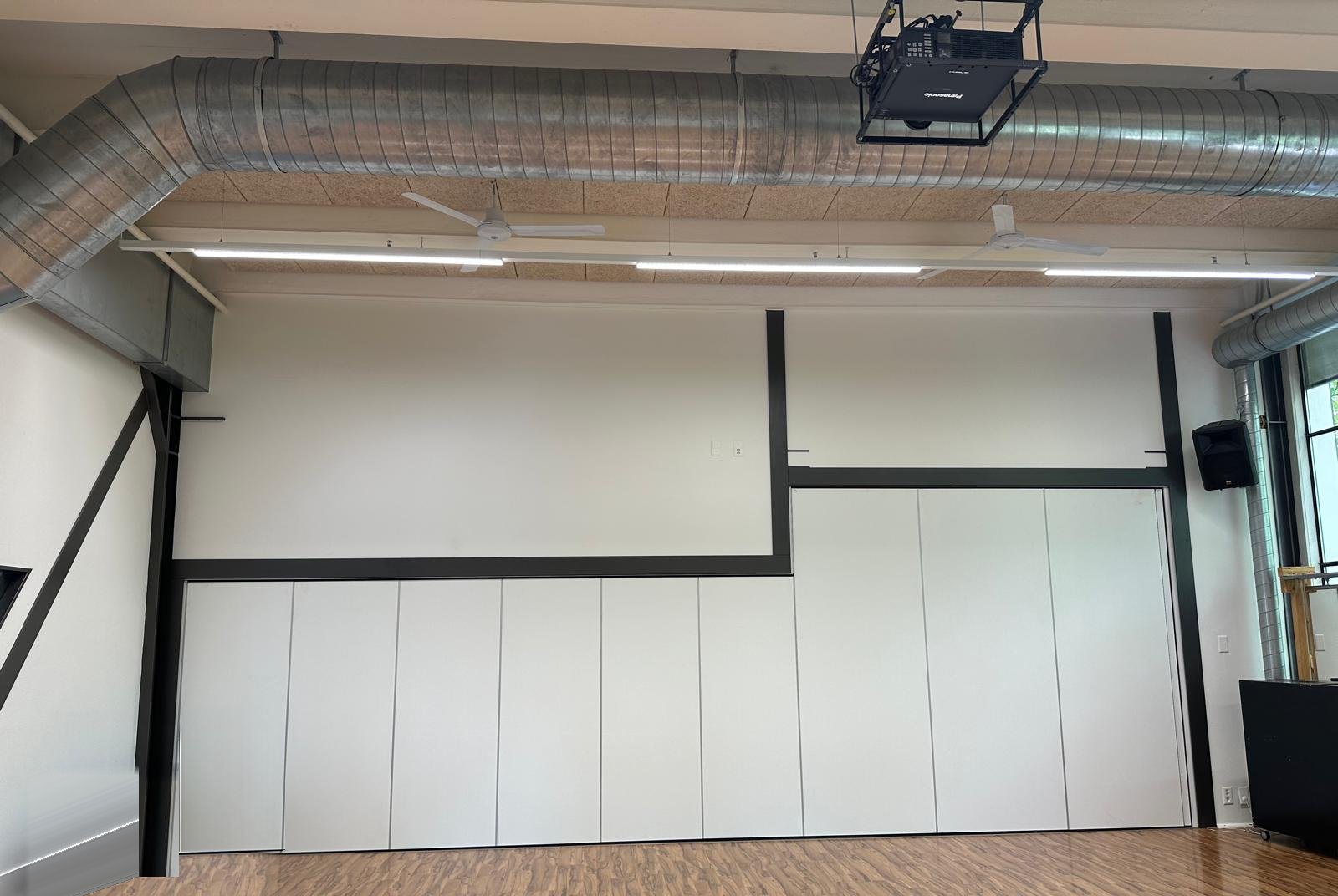 Leere Stirnwand im Workshopraum mit Schiebewand und schwarzem Stahlrahmen, an der Decke der bisherige Beamer